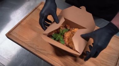 Şef aşçıya yakın çekim. Ellerinde tıbbi eldivenlerle karton konteynırda yiyecek paketliyor.