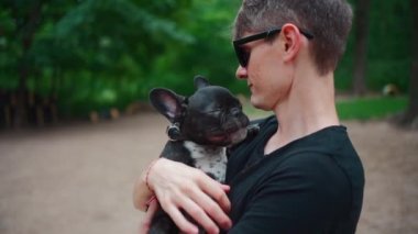 Yakışıklı erkek, yaz parkında sevimli bir köpek yavrusuyla ya da buldogla sarılıp oynuyor.