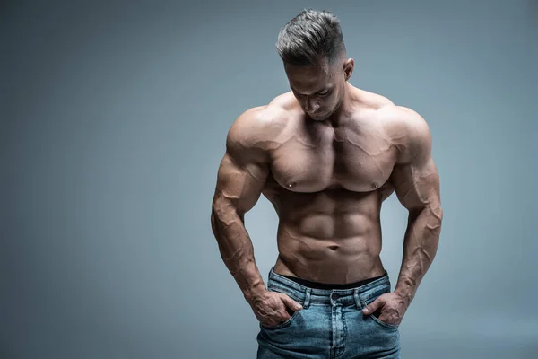 Jovem atleta bonito fisiculturista posando em jeans no fundo cinza — Fotografia de Stock