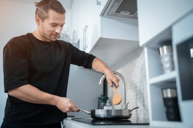Modern ev mutfağında ahşap kaşıkla yemek pişiren çekici adam gülümsüyor.
