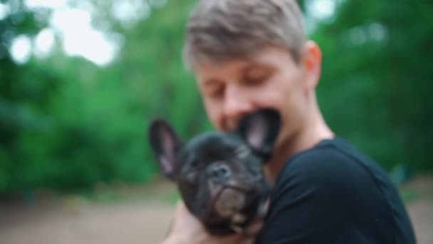 En kjekk mann som klemmer og leker med en søt valp eller fransk bulldogg i sommerparken – stockvideo