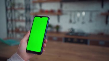 Ev mutfağında yeşil ekran modeli olan akıllı telefon kullanarak kadın elinin yakın plan görüntüsü.