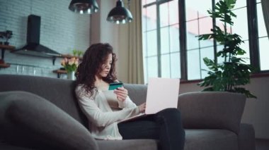 Genç bir kadın evdeki koltukta kredi kartı ve dizüstü bilgisayarla online alışveriş yapıyor.