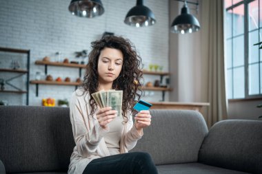 Genç, esmer bir kadın evdeki kanepede nakit veya kredi kartı seçiyor.