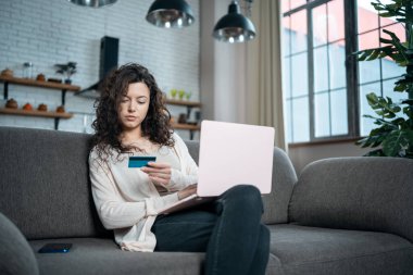 Genç bir kadın evdeki koltukta kredi kartı ve dizüstü bilgisayarla online alışveriş yapıyor.