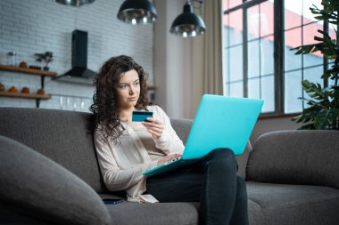 Genç bir kadın evdeki koltukta kredi kartı ve dizüstü bilgisayarla online alışveriş yapıyor.
