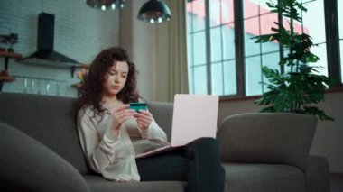 Genç bir kadın evdeki koltukta kredi kartı ve dizüstü bilgisayarla online alışveriş yapıyor.