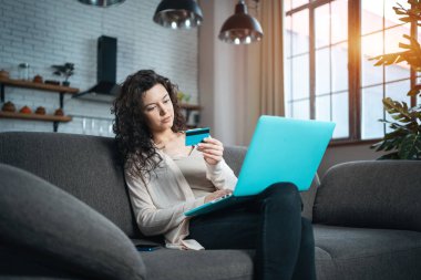 Genç bir kadın evdeki koltukta kredi kartı ve dizüstü bilgisayarla online alışveriş yapıyor.