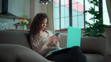 Genç bir kadın evdeki koltukta kredi kartı ve dizüstü bilgisayarla online alışveriş yapıyor.