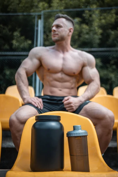 Πρωτεΐνη ορού γάλακτος σε σκόνη σε μαύρο δοχείο και αθλητικός άνθρωπος κάθεται σε εξωτερικούς χώρους — Φωτογραφία Αρχείου