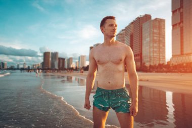 Güneşli günbatımında sahilde duran kaslı vücutlu yakışıklı beyaz adam, denizde ya da okyanus kıyısında yürüyen çekici erkek atlet. Yüksek kalite fotoğraf
