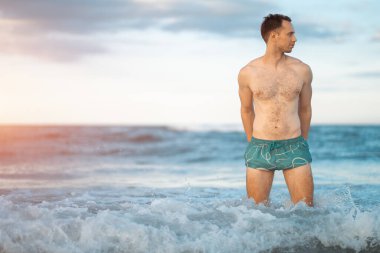 Güneşli günbatımında sahilde duran kaslı vücutlu yakışıklı beyaz adam, denizde ya da okyanus kıyısında yürüyen çekici erkek atlet. Yüksek kalite fotoğraf