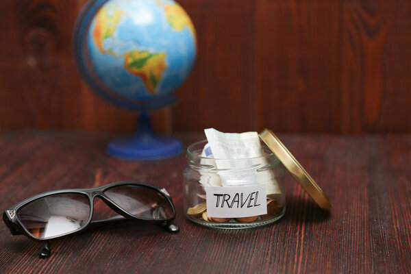 Collecting money for travel. Glass jar as moneybox with cash savings, paper label on wooden table. Sunglasses, and globe at background