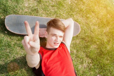 Young man lying on grass shows peace sign, skateboard near