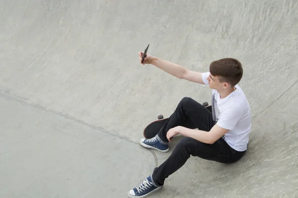 Młody człowiek siedzi w pobliżu planszę skate za pomocą smartfonu — Zdjęcie stockowe