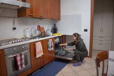 Mutfakta çömelmiş bir kadın, kirli bulaşıkları, çatal bıçakları ve bardakları açık bir bulaşık makinesine yerleştiriyor, ev işleri ve günlük rutin işlerle meşgul.