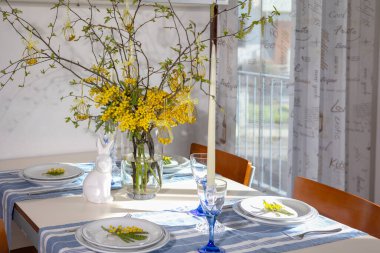 Paskalya kutlaması için dekore edilmiş yemek masası. İçinde mimoza dalları olan bir süs. Süslü paskalya yumurtası süsü. Seramik bir tavşan. Ve mavi çizgili koşucularla yer ayarları