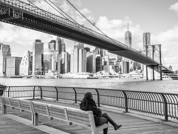 A woman below the Brooklyn Bridge watching the Manhattan skyline.