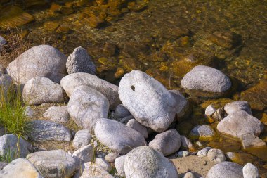 Temiz ve sığ suyun kenarında uzanan pürüzsüz nehir taşları, şeffaf dereden yansıyan güneş ışığıyla doğal bir nehir kenarı manzarası yaratıyor..