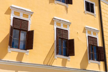 Üç klasik penceresi ve güneş ışığına gölge düşüren ahşap panjurları olan tarihi sarı bina. Dekoratif beyaz çerçeveler geleneksel Avrupa mimari ayrıntılarını ve zarif simetriyi vurguluyor.