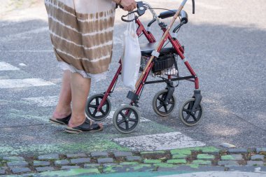 Kıdemli bir kadın gündüz vakti bir tekerlekli sandalyeyle karşıdan karşıya geçiyor. Kentsel yaşamda bağımsızlık, ulaşılabilirlik ve aktif yaşlanmayı gösteren günlük hareketlilik yardım sahnesi.