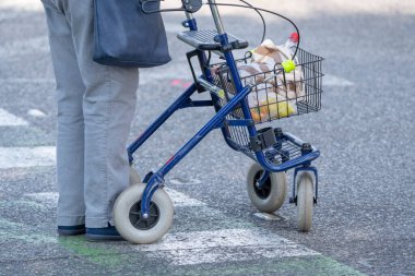 Yaşlı kişi, şehir caddesinde bakkaliye malzemelerini taşıyan bir tekerlekli sandalyeyle ayakta duruyor. Bağımsızlığı, ulaşılabilirliği ve açık havada aktif günlük yaşamı gösteren günlük gezici yardım sahnesi.