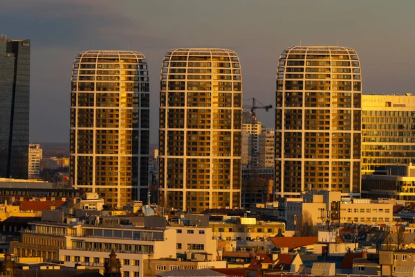 Sıcak bir akşam ışığında şehir manzarasının üzerinde yükselen üç tane modern gökdelenli yerleşim kulesi. Yuvarlak cepheli, yoğun gelişimli ve arka planda inşaat vinci olan çağdaş mimari sahnesi.