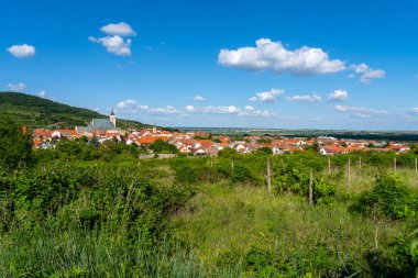 Kırmızı çatıları ve kilise kuleleri olan bir köyün panoramik manzarası. Parlak mavi gökyüzünün altında, dağınık bulutlarla çevrili yeşil üzüm bağlarıyla çevrili..