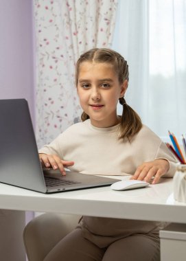 Liseli kız dizüstü bilgisayarla evde oturuyor, uzaktan çalışıyor. Uzaktan öğrenme, online eğitim konsepti. Çocuk ev ortamında, gün ışığında masa başında ödev yapıyor..