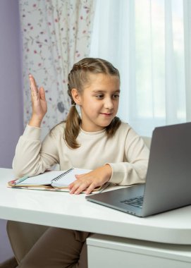 Gülümseyen kız evde online derse katılırken elini kaldırıyor. Masada dizüstü bilgisayarla oturuyor. Aktif öğrenim kavramı, sanal eğitim, öğrenci katılımı.