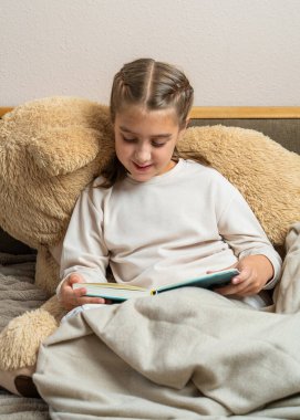 Yatakta oturup kitap okuyan tatlı bir kız, büyük bir oyuncak ayının karşısında dinleniyor. Çocukluk, rahatlık, öğrenme, aile hayatı.