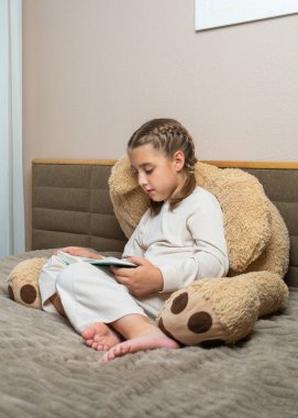 Küçük tatlı kız yatağında dev bir oyuncak ayıya yaslanırken kitap okuyor. Samimi ve huzurlu bir an çocukluk, hayal gücü, erken öğrenme, ev konforu.