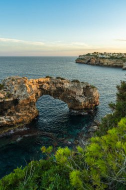Cala Santany 'deki kayalık deniz kemerinin gün batımı manzarası. Sıcak ışık, Akdeniz kıyılarının sakin sularını ve doğal uçurumlarını yansıtıyor.