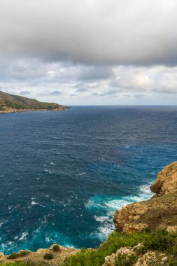 İspanya, Mallorca 'daki kayalık kıyı şeridi canlı mavi deniz ve bulutlu gökyüzü. Akdeniz kıyı manzarası, doğa güzelliği, seyahat için mükemmel, tatil görüntüleri..