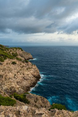 Far de Capdepera, Mallorca yakınlarındaki gri fırtına bulutları altında kayalık kıyı şeridine çarpan dalgaların dramatik görüntüsü. Doğa gücü kavramı, deniz manzarası, engebeli sahil..