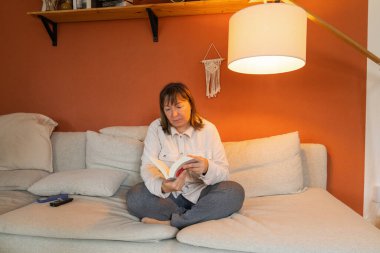 Orta yaşlı bir kadın kanepede oturuyor, telefonunu bir kenara bırakıp kitap okuyor. Yer lambası yanıyor, rahatlatıcı bir atmosfer yaratıyor. Rahatlama, sakin bir akşam havası, huzurlu bir ev hayatı..