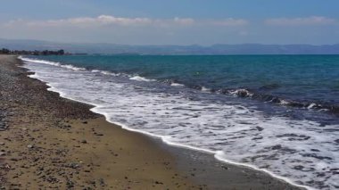 Kıbrıs 'ın güney kıyısındaki çakıl taşlı plajda deniz dalgaları yavaşça yuvarlanıyor. Doğal deniz manzarası, huzurlu kıyı atmosferi, yaz yolculuğu, sakin bir arka plan..