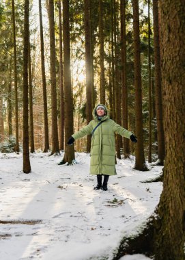 Kış ormanında kollarını açmış, yukarı bakıp gülümseyen, sıcak bir palto giyen mutlu genç kadın. Güneş ışığı, karlı ağaçlar neşeli, büyülü kış atmosferi yaratır..