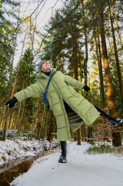 Karlı kış ormanlarında kolları açık bir şekilde tek bacağının üzerinde dengede duran, sıcak bir palto giymiş neşeli genç kadın. O mutlu bir şekilde güler. Neşeli, büyülü kış havası.