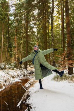 Kış yastıklı palto giyen neşeli genç bir kadın orman manzarasının tadını çıkarıyor. Kolları açık bir şekilde tek bacağının üzerinde duruyor, yukarı bakıyor ve gülüyor..