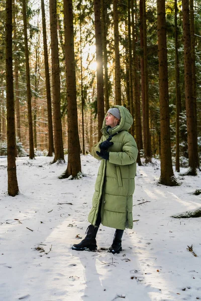 Kış ormanında dikilmiş, sıcak bir palto giyen sevimli genç bir kadın, yana bakıyor. Güneş ışınları karlı ağaçların arasında parlıyor. Akıl sağlığı, sakin yürüyüş, kış doğası, rahat yaşam tarzı..
