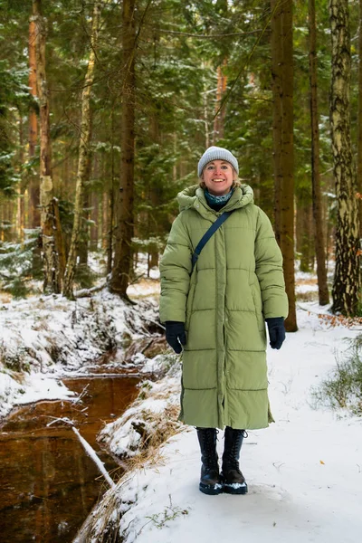 Sakin orman manzarasında kışlık ceketini giyen huzurlu genç bir kadın. Güneş ışınları karla kaplı ağaçların arasından geçer, gevşemeyi sembolize eder, kışın yavaş yaşamayı..