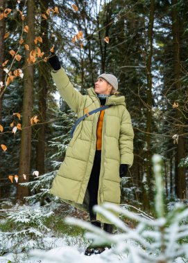 Sakin orman manzarasında kışlık ceketini giyen huzurlu genç bir kadın. Güneş ışınları karla kaplı ağaçların arasından geçer, gevşemeyi sembolize eder, kışın yavaş yaşamayı..