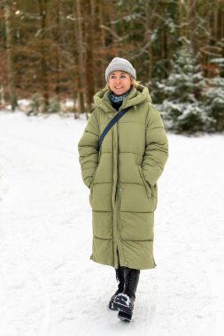 Young woman in warm outerwear standing in winter forest environment. Gentle sun rays and snow covered trees create calm, natural winter mood.