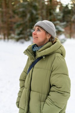 Sıcak giysili genç bir kadın kış ormanı ortamında dikiliyor. Yumuşak güneş ışınları ve karla kaplı ağaçlar sakin, doğal kış havası yaratır..
