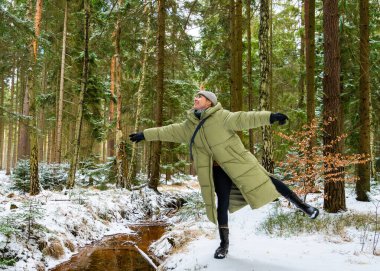 Yastıklı ceketli gülümseyen genç kadın orman manzarasında eğleniyor. Kolları açık bir şekilde tek ayak üzerinde duruyor ve yukarı bakıyor, doğal olarak gülüyor, mutluluğu temsil ediyor..