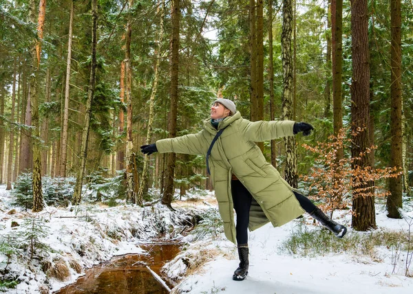 Yastıklı ceketli gülümseyen genç kadın orman manzarasında eğleniyor. Kolları açık bir şekilde tek ayak üzerinde duruyor ve yukarı bakıyor, doğal olarak gülüyor, mutluluğu temsil ediyor..