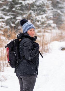 Kar fırtınasında sırt çantasıyla gezen orta yaşlı bir kadın arkasına dönüp kameraya bakıyor. Görüş mesafesi düşük ve kış koşulları macera ve kararlılığı simgeliyor..