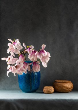 Manolya çiçekleri gri arkaplandaki mavi vazoya yerleştirilmiş, yanına da iki hasır kutu yerleştirilmiş. Uzayı kopyala Zarif çiçekli natürmort, minimalist ev dekorasyonu.