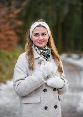 Sıcak kış kıyafetleri içinde neşeli genç bir kadın dışarıda soğuk bir günün tadını çıkarıyor. Palto, eldiven ve örülmüş saç bandı takan gülümseyen bir kadın karlı bir ormanda eğleniyor..